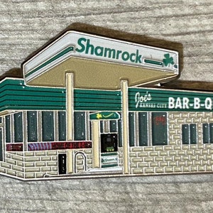 May include: A green and white enamel pin depicting a vintage gas station with the text "Shamrock" and "Joe's Kansas City BAR-B-QUE" on the front.