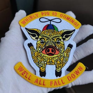 May include: Embroidered patch featuring a stylized pig with intricate yellow patterns, wearing a colorful hat. The patch has the text "WHIRLY PIG IS COMING!" and "WELL ALL FALL DOWN" in yellow on a white and yellow background.
