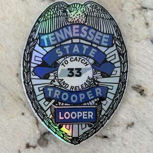 May include: A 3-inch holographic sticker of a Tennessee State Trooper badge. The badge is blue and silver with the words "Tennessee State" and "Trooper" on it. The number "33" is also on the badge, along with the words "To Catch and Release". The bottom of the badge says "Looper".