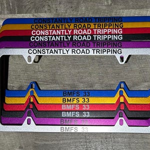 May include: A stack of license plate frames in various colors, including blue, gold, red, black, and purple. Each frame has the text "CONSTANTLY ROAD TRIPPING" and "BMFS 33". The frames are made of metal.