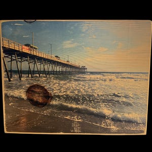 May include: Wooden wall art featuring a scenic view of a pier extending into the ocean. The image captures the pier's structure, the waves, and the sky with a warm, vintage aesthetic. The artwork is mounted on a wooden plank.