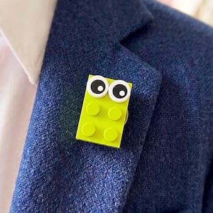 May include: A lime green rectangular building block pin attached to a navy blue textured blazer. The pin features two white circular eyes with black pupils positioned at the top, giving the block a character-like appearance on the lapel.
