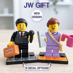 May include: Two custom minifigures labeled JW GIFT and NEW DESIGN. The left figure wears a black pinstripe suit, holding a black microphone and an orange Teaching Toolbox case. The right figure wears a lavender dress, holding a Holy Scriptures book and a pink mug. Both stand on black bases labeled Best Life Ever.