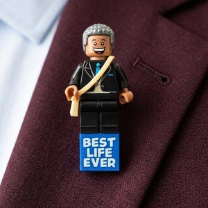 May include: A lapel pin featuring a plastic minifigure with gray hair, a black suit, and a blue tie. The figure wears a tan sash and stands atop a blue rectangular base with the white text BEST LIFE EVER printed on the front surface.
