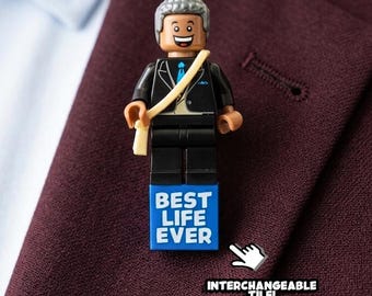 Puede incluir: Un pin de solapa con una minifigura de plástico de pelo gris, traje negro y corbata azul. La figura lleva una banda de color beige y se apoya sobre una base rectangular azul con el texto blanco BEST LIFE EVER. Un gráfico indica una pieza intercambiable.