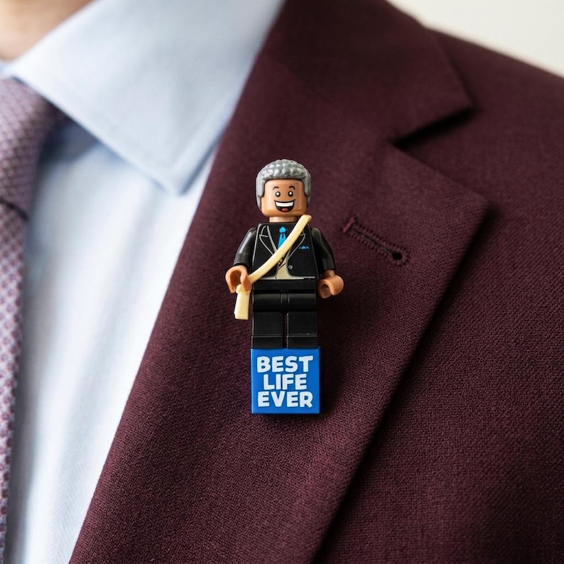 May include: A lapel pin featuring a plastic minifigure with gray hair, a black suit, and a blue tie. The figure wears a tan sash and stands atop a blue rectangular base with the white text BEST LIFE EVER printed on the front surface.