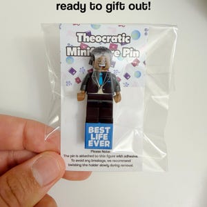 May include: A plastic minifigure pin featuring a smiling character in a black suit with a blue tie, mounted on a white display card inside a clear plastic bag. The card displays the text Theocratic Miniature Pin and Best Life Ever.