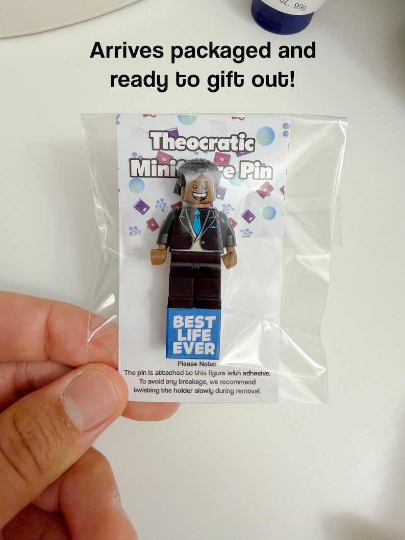 May include: A plastic minifigure pin featuring a smiling character in a black suit with a blue tie, mounted on a white display card inside a clear plastic bag. The card displays the text Theocratic Miniature Pin and Best Life Ever.