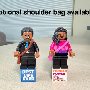 May include: Two plastic minifigures standing on rectangular blocks. The left figure wears a black suit with a blue tie and a brown shoulder bag. The right figure wears a pink top and a pink shoulder bag. Text reads: Optional shoulder bag available!