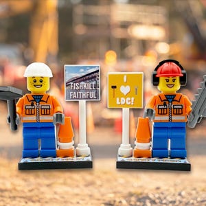May include: Two construction worker minifigures wearing orange vests, blue pants, and hard hats. One holds a hammer, the other a chainsaw. They stand on black bases labeled SAFETY FIRST next to signs reading FISHKILL FAITHFUL and I HEART LDC.