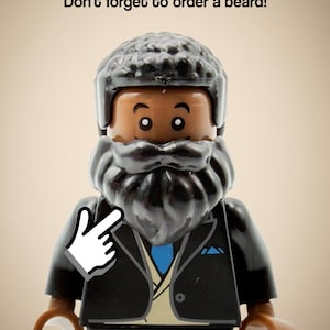 May include: A plastic minifigure with dark brown skin, a textured black hair piece, and a thick black beard. The figure wears a black suit jacket over a tan vest and a blue tie. Above the figure, the text reads: Don't forget to order a beard!