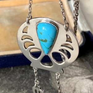 May include: A silver-toned pendant necklace featuring a central triangular turquoise stone with green veining. The pendant has a cut-out fan-like design and two small dangling heart-shaped charms attached to the base by thin silver-toned chains.