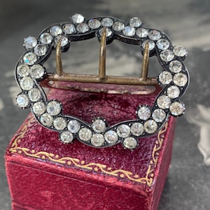 May include: An antique silver-tone buckle with clear, round rhinestones set in a floral pattern. The buckle has three prongs and is displayed on a red velvet box, showcasing its vintage design.