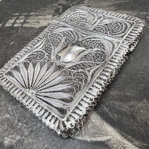 May include: A rectangular silver filigree box with detailed floral and geometric patterns. The box has a central shield-shaped element and a scalloped edge. The intricate metalwork creates a delicate and ornate appearance, showcasing craftsmanship.
