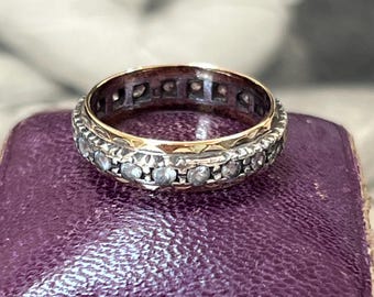 Puede incluir: Un anillo de banda bicolor que presenta una sección central de color plateado engastada con una hilera continua de pequeñas piedras claras, flanqueada por bordes lisos de color dorado. La banda metálica muestra un acabado texturizado.