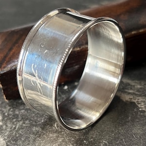 Könnte beinhalten: Ein polierter silberner Serviettenring mit eingravierten Initialen. Der Ring hat ein breites Band mit einem Perlenrand und einer glatten, reflektierenden Oberfläche. Die Initialen sind in einer zarten Schrift. Ein klassisches Stück Tischdekoration.