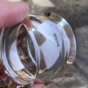 May include: A close-up of a shiny, silver napkin ring. The ring has a rounded shape with a smooth, reflective surface. The hallmarks are visible on the surface. The ring is held by a person's hand.