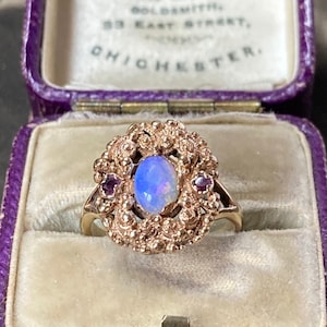 May include: A rose gold ring with an oval, opalescent blue gemstone at the center, surrounded by intricate filigree and two smaller, round, red gemstones. The ring is displayed in a purple velvet lined box.