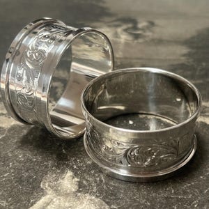 May include: Two polished silver napkin rings with an engraved floral pattern. The rings are circular and have a bright, reflective surface. The napkin rings are displayed on a dark, textured surface, creating a contrast.