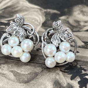 以下が含まれることがあります： それぞれ6つの模造真珠が付いた、銀色のイヤリングのペア。イヤリングは葉のデザインと湾曲したフレームが特徴です。真珠は白く、虹色の光沢があります。背景はヴィンテージで、ジュエリーに焦点を当てています。