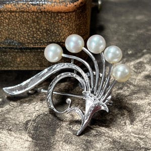 Könnte beinhalten: Silberfarbene Brosche mit fünf runden, weißen Perlen in floralem Design. Die Brosche hat aufwendige Details, darunter ein geschwungenes, strukturiertes Element und eine vasenartige Basis. Der Hintergrund ist eine strukturierte, neutrale Oberfläche.