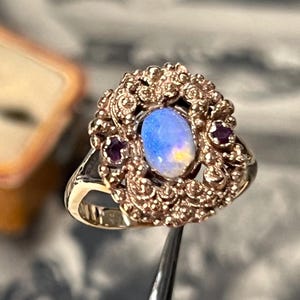 Puede incluir: Un anillo de oro con una ópalo ovalado con tonos azules y amarillos, rodeado por un engaste detallado y ornamentado. Dos pequeñas gemas de color rojo oscuro flanquean el ópalo. El anillo es sujetado por unas pinzas.