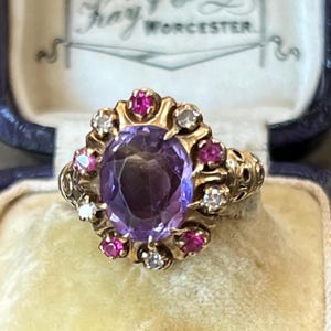 May include: A gold ring featuring a large oval-cut purple amethyst center stone surrounded by a halo of alternating small round-cut pink rubies and clear white diamonds. The metal band has an ornate, textured design along the sides.