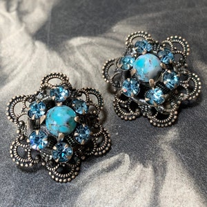May include: Pair of vintage clip-on earrings. Each earring features a central turquoise-colored stone surrounded by smaller blue crystals. The stones are set in a filigree metal flower design. The earrings have a classic, elegant style.