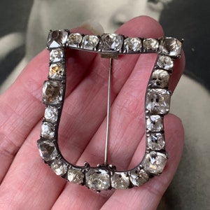 May include: A U-shaped buckle brooch featuring a dark metal frame set with numerous clear, faceted, square-cut stones. The piece includes a thin, straight metal pin mechanism attached to the back, designed to secure the buckle to fabric.