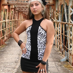 May include: A woman wearing a black and white patterned mini dress with a halter neckline, standing on a rusty bridge. She is wearing white platform sneakers and has her hair pulled back.
