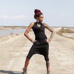 Puede incluir: Una mujer con trenzas rojas lleva un mono negro sin mangas con cuello de pico y se encuentra de pie en un camino de tierra. Lleva sandalias de gladiador con hebillas.