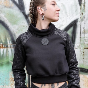 Op de afbeelding: Zwart cropped sweatshirt met een hoge kraag en mouwen met bloemenpatroon. Een cirkelvormig embleem is gecentreerd op de borst. Het model draagt een zwarte jeans en heeft meerdere piercings en tatoeages.