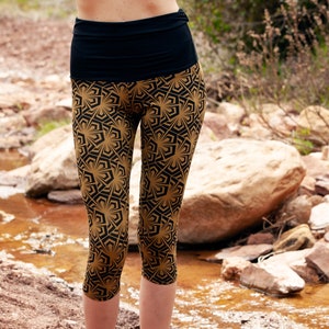 May include: A pair of gold and black patterned leggings with a black waistband. The leggings are cropped and the model is wearing black sandals with gold ankle bracelets.