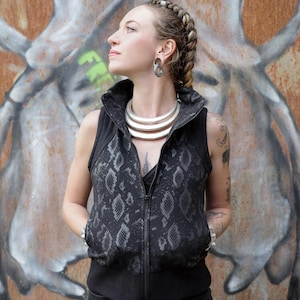 May include: A black vest with a snakeskin pattern on the front, featuring a high collar and a full-length zipper. The person is wearing a silver necklace and earrings. The background is a graffiti-covered wall.