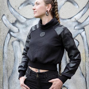 Pode incluir: Camisola preta curta com gola alta e detalhes estampados nos ombros e mangas. A camisola apresenta um emblema redondo no peito. A pessoa na imagem está usando jeans pretos e vários anéis.