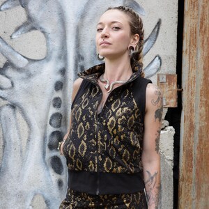 May include: A black and gold snakeskin print vest and matching skirt. The vest has a high collar and a zipper closure. The skirt is a mini skirt. The outfit is worn with fishnet stockings and jewelry.