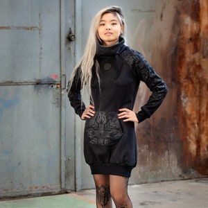 Puede incluir: Un vestido negro de cuello alto con un estampado floral en la parte delantera y las mangas. El vestido tiene un bolsillo canguro en la parte delantera. La modelo lleva medias de rejilla negras y botines negros.