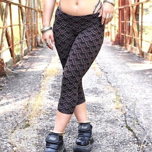 May include: Black and brown patterned capri leggings. The leggings feature a repeating geometric design. The person is wearing black platform boots with gold accents. The background is a weathered metal bridge.