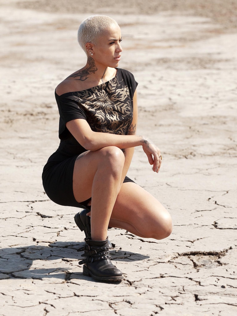 May include: A woman with short, blonde hair is wearing a black off-the-shoulder top with a gold floral pattern. She is wearing black boots and is squatting on a cracked, dry surface.