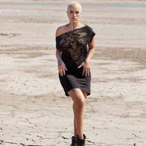 May include: A woman wearing a black off-the-shoulder dress with a gold floral design. She is standing in a desert landscape with cracked earth.