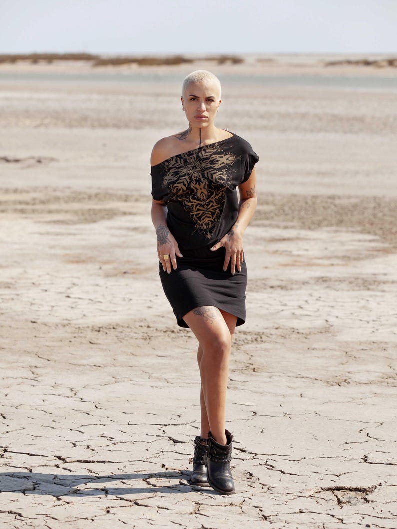 May include: A woman wearing a black off-the-shoulder dress with a gold floral design. She is standing in a desert landscape with cracked earth.