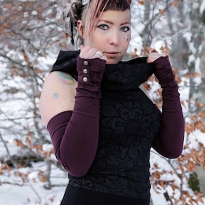 May include: A woman with pink and white hair wearing a black, high-necked top with a lace pattern and long, burgundy sleeves. She is wearing a silver ring on her finger and has a tattoo on her arm.