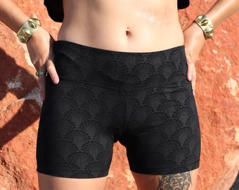 May include: Black athletic shorts with a textured, repeating pattern. The shorts are high-waisted and form-fitting. The model is wearing a black crop top and gold bracelets. The background is a reddish-brown color.