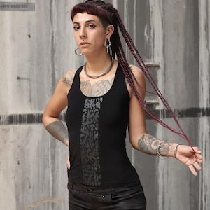 Puede incluir: Una mujer con el pelo rojo trenzado lleva un top negro de tirantes con un diseño abstracto gris y pantalones cortos de mezclilla negros. Tiene varios tatuajes en los brazos y las piernas.