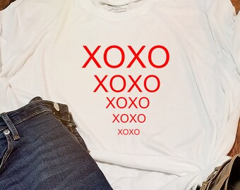 Könnte beinhalten: Ein lässiges Outfit mit einem weißen T-Shirt mit rotem "XOXO"-Text, dunkelblauen Jeans und hellbraunen Stiefeletten. Die Artikel sind auf einer Holzoberfläche angeordnet, was auf ein Konfektionsensemble hindeutet.