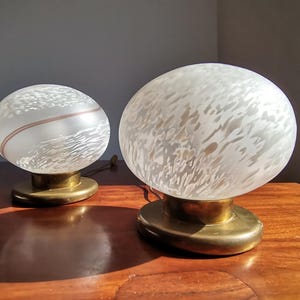 May include: Two vintage table lamps with frosted glass globes and brass bases. One lamp has a brown stripe around the globe. The lamps are on a wooden surface. The lamps are round and have a mid-century modern design.