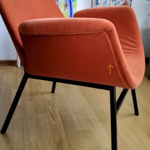 May include: A burnt orange armchair with black metal legs. The chair has a curved back and a slightly angled seat. The chair is sitting on a wooden floor.