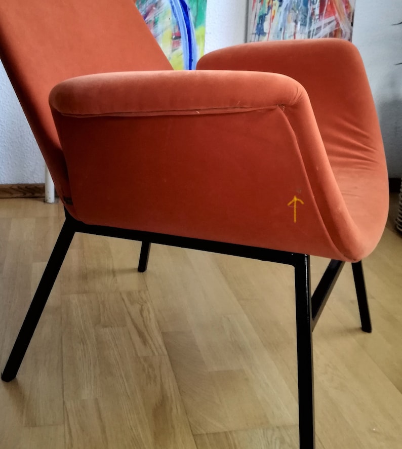 May include: A burnt orange armchair with black metal legs. The chair has a curved back and a slightly angled seat. The chair is sitting on a wooden floor.