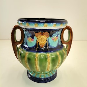 Può includere: Un vaso in ceramica blu e verde con due manici marroni. Il vaso ha un design decorativo con un viso grottesco e motivi floreali.