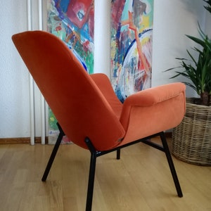 May include: A bright orange velvet armchair with black metal legs. The chair is sitting on a wooden floor.
